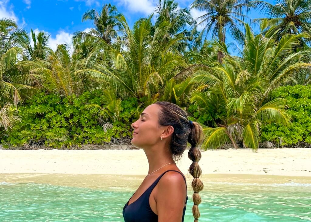 metida en el agua hasta la cintura en el mar del océano indico y de fondo una isla local llena de vegetación, palmeras verdes, agua turquesa